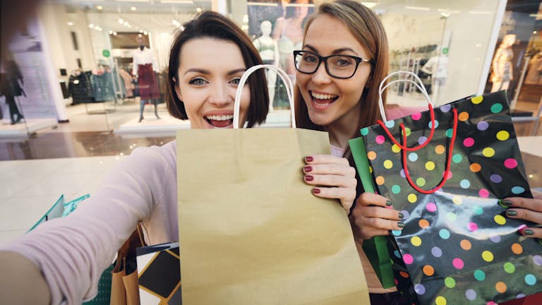 Two women smiling with shopping bags in a mall environment.